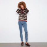 A model with curly brown hair stands against a white wall wearing a beige leopard-print hoodie with red trim, blue skinny jeans, and brown loafers.