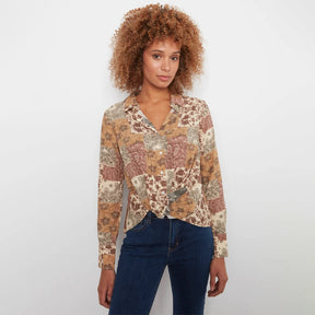 A woman with curly light brown hair wearing a long-sleeve button-up shirt with a multicolor patchwork floral pattern in beige, brown, and rust tones, paired with dark blue jeans, standing against a plain white background.