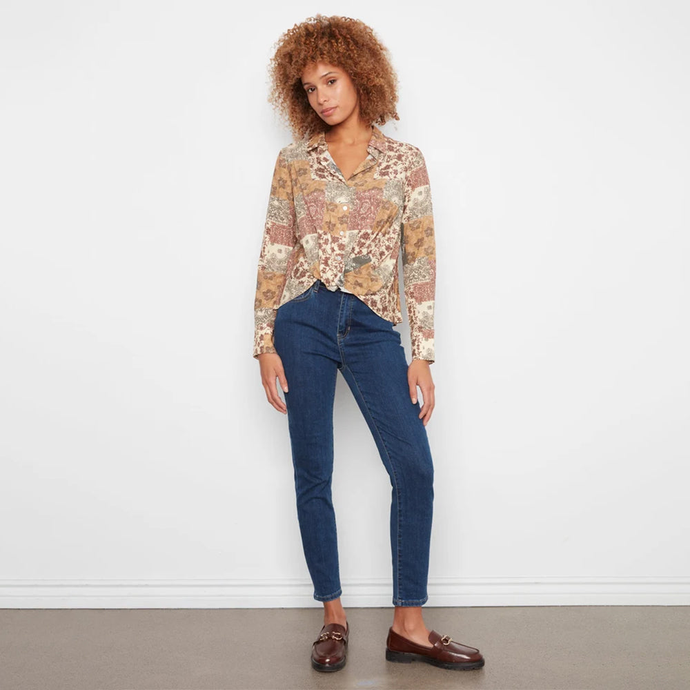 A woman with curly brown hair stands against a white wall wearing a long-sleeve button-up blouse with a beige, brown, and gold floral pattern, dark blue skinny jeans, and brown leather loafers.
