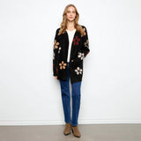 A woman stands against a plain white wall wearing a black open-front cardigan with large multicolor floral motifs, a white top, blue jeans, and brown ankle boots.