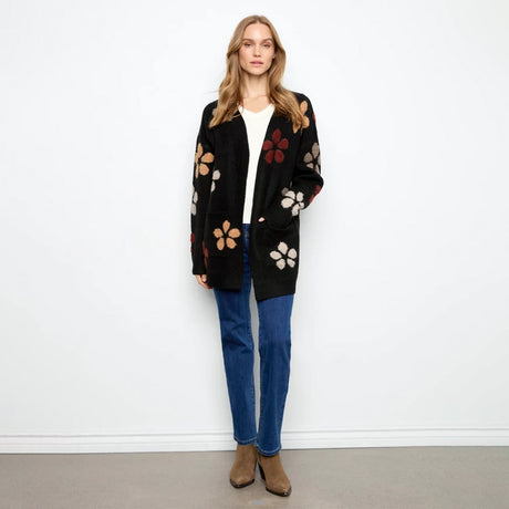A woman stands against a plain white wall wearing a black open-front cardigan with large multicolor floral motifs, a white top, blue jeans, and brown ankle boots.