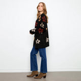 A woman stands in profile wearing a black knit cardigan with large floral motifs in red, beige, and brown, paired with blue jeans and brown ankle boots, against a plain white wall.