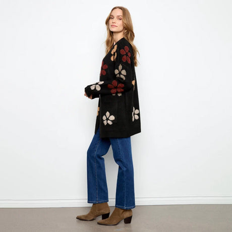 A woman stands in profile wearing a black knit cardigan with large floral motifs in red, beige, and brown, paired with blue jeans and brown ankle boots, against a plain white wall.