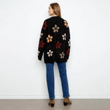 A woman stands with her back to the camera wearing a black knit cardigan with large textured floral motifs in beige, rust red, and brown, paired with blue jeans and brown ankle boots, against a plain white wall.