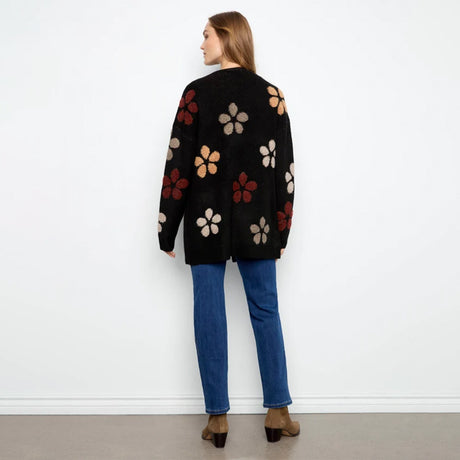 A woman stands with her back to the camera wearing a black knit cardigan with large textured floral motifs in beige, rust red, and brown, paired with blue jeans and brown ankle boots, against a plain white wall.