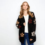 A woman modeling a black open-front cardigan with large multicolor floral motifs in red, beige, and brown, worn over a white top and blue jeans against a plain white background.