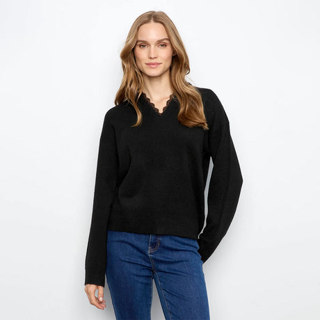 A woman modeling a black long-sleeve knit sweater with a scalloped V-neckline, paired with blue jeans, against a plain white studio background.