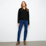 A woman stands against a plain white wall wearing a black long-sleeve top with a scalloped V-neckline, blue jeans, and brown ankle boots.