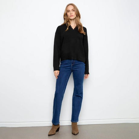 A woman stands against a plain white wall wearing a black long-sleeve top with a scalloped V-neckline, blue jeans, and brown ankle boots.