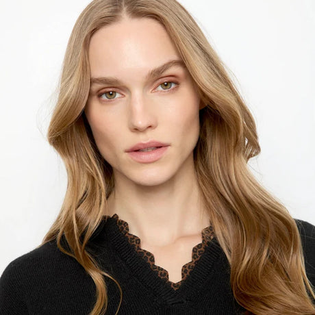 A close-up portrait of a woman with long wavy light brown hair wearing a black knit sweater with a brown leopard-print scalloped V-neck trim, set against a plain white background.
