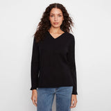 A woman with long, curly brown hair wearing a black long-sleeve V-neck knit top and blue jeans, standing against a plain white studio background.