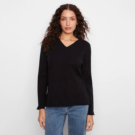 A woman with long, curly brown hair wearing a black long-sleeve V-neck knit top and blue jeans, standing against a plain white studio background.