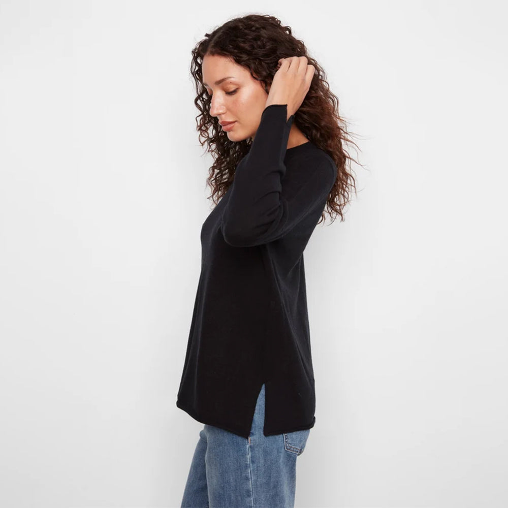 A woman with curly brown hair wearing a black long-sleeve top and blue jeans, shown from a side profile against a plain white studio background.
