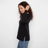 A woman with curly brown hair wearing a black long-sleeve top and blue jeans, shown from a side profile against a plain white studio background.