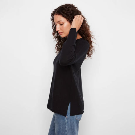 A woman with curly brown hair wearing a black long-sleeve top and blue jeans, shown from a side profile against a plain white studio background.