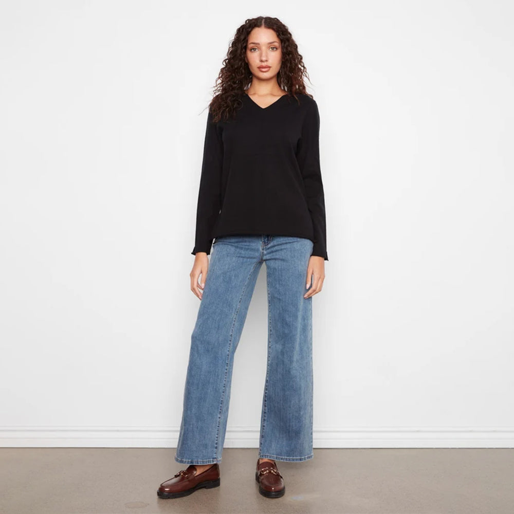 A woman stands against a plain white wall wearing a black long-sleeve V-neck top, wide-leg blue jeans, and brown leather loafers.