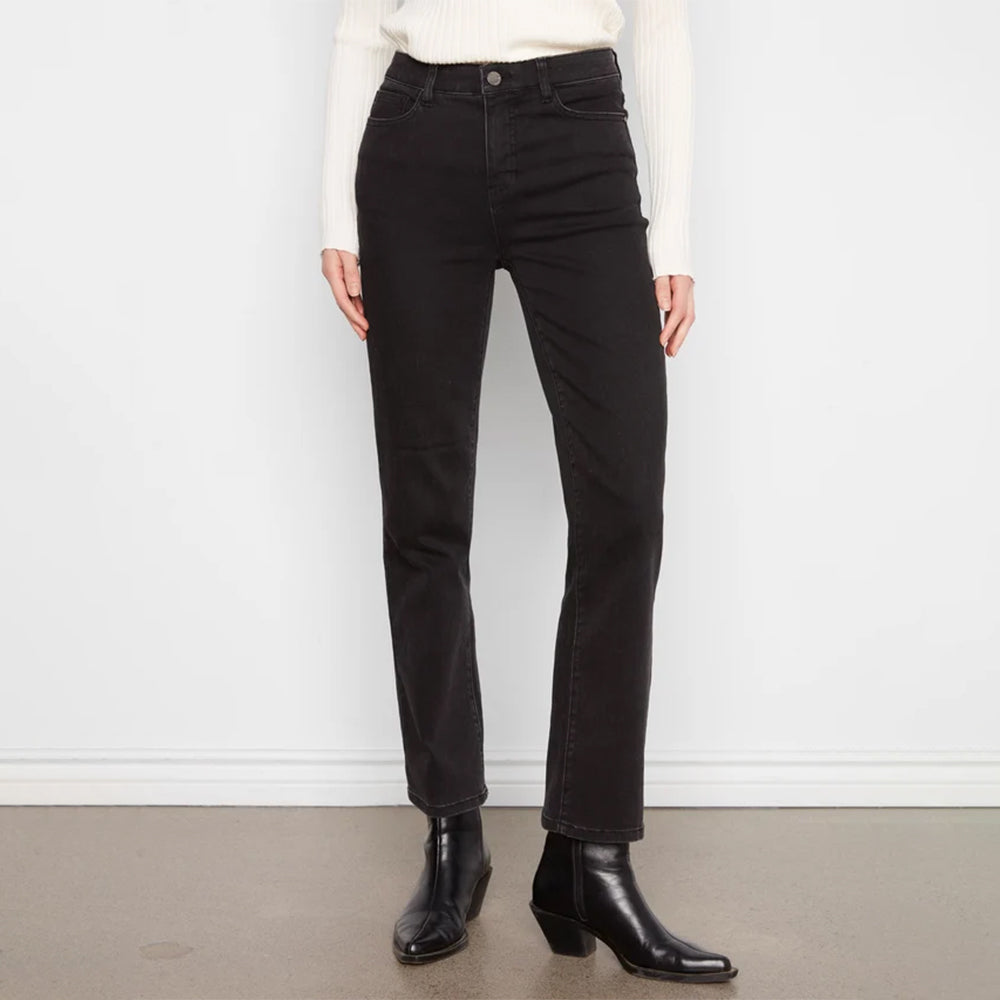 A person wearing black straight-leg jeans, a white ribbed sweater, and black ankle boots, standing against a white wall and gray floor.