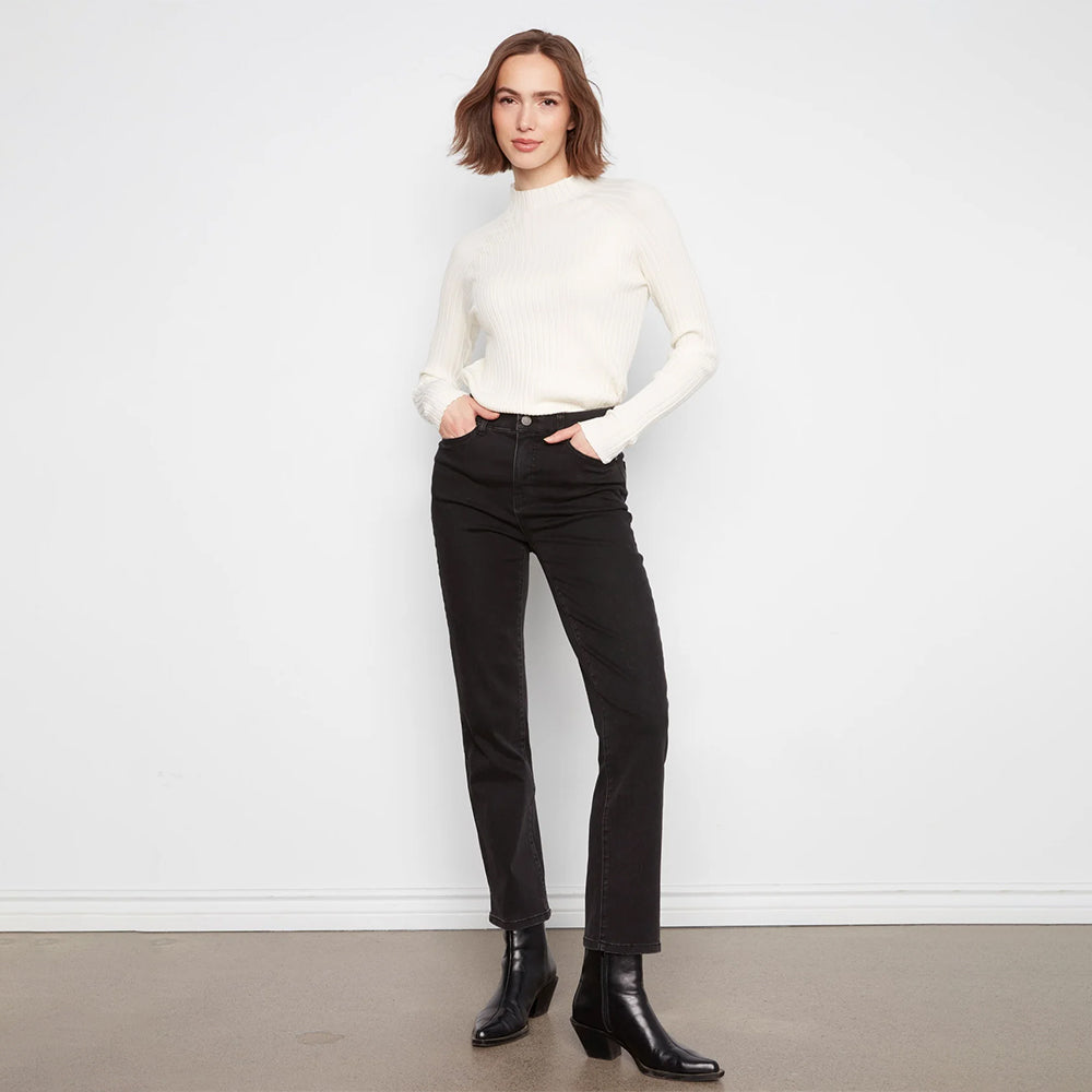 A woman stands against a white wall wearing a fitted off-white ribbed mock-neck sweater, black straight-leg jeans, and black ankle boots.