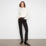 A woman stands against a white wall wearing a fitted off-white ribbed mock-neck sweater, black straight-leg jeans, and black ankle boots.