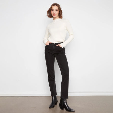A woman stands against a white wall wearing a fitted off-white ribbed mock-neck sweater, black straight-leg jeans, and black ankle boots.