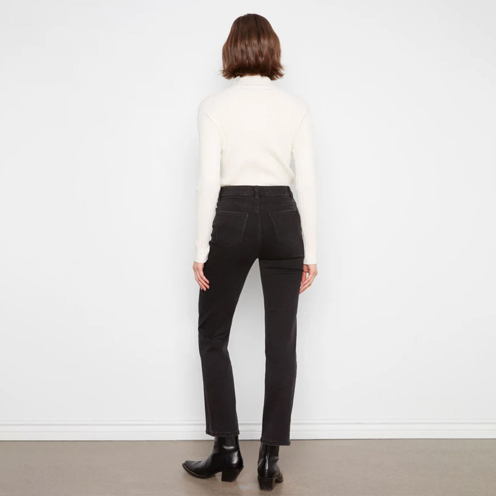A model viewed from the back wearing a cream ribbed turtleneck sweater, black high-waisted straight-leg jeans, and black leather ankle boots, standing against a white wall.