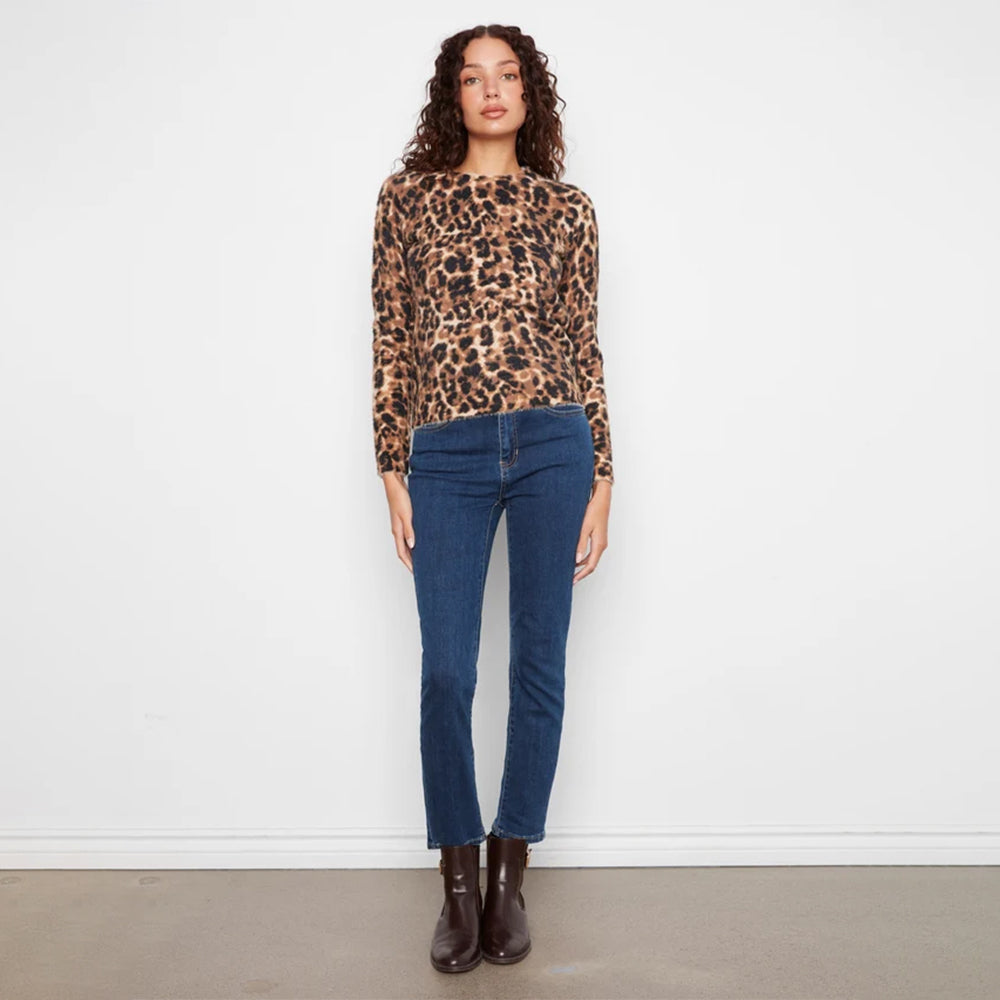 A woman stands against a plain white wall wearing a leopard-print long-sleeve top, dark blue jeans, and dark brown ankle boots.