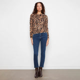 A woman stands against a plain white wall wearing a leopard-print long-sleeve top, dark blue jeans, and dark brown ankle boots.