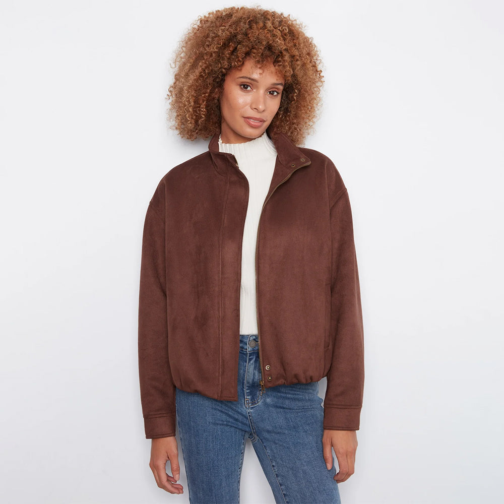 A woman with curly brown hair wearing a brown suede jacket over a white turtleneck and blue jeans, standing against a plain white background.