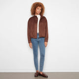 A woman with curly light brown hair stands against a white wall wearing a brown suede-like cropped jacket over a white top, blue skinny jeans, and brown loafers.