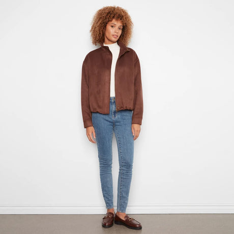 A woman with curly light brown hair stands against a white wall wearing a brown suede-like cropped jacket over a white top, blue skinny jeans, and brown loafers.