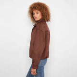 A woman with curly brown hair wearing a brown suede jacket and blue jeans, shown from a side profile against a plain white studio background.