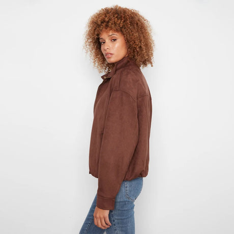 A woman with curly brown hair wearing a brown suede jacket and blue jeans, shown from a side profile against a plain white studio background.