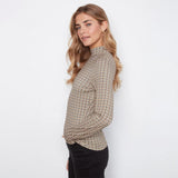 A woman wearing a beige and brown checkered mock neck long-sleeve top with black pants, standing against a plain white background.