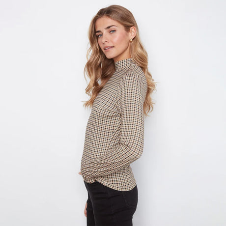 A woman wearing a beige and brown checkered mock neck long-sleeve top with black pants, standing against a plain white background.