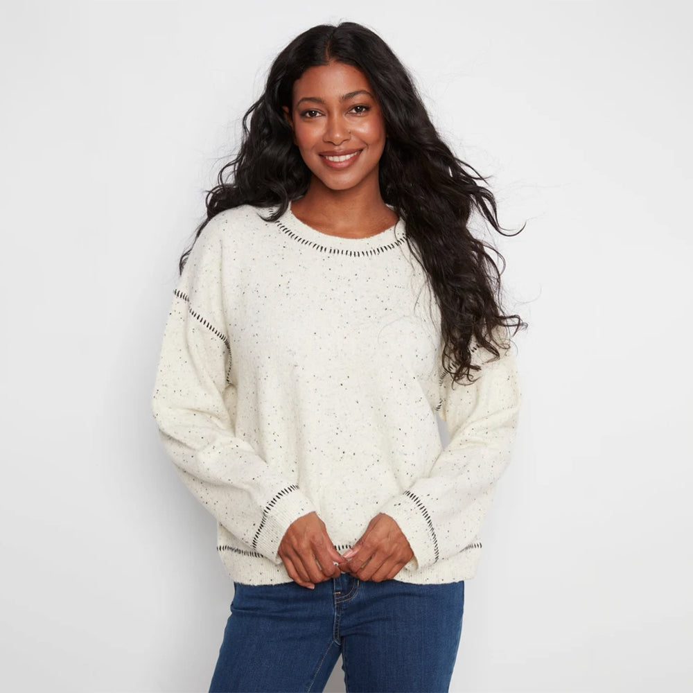 A woman wearing a cream-colored knit sweater with small black speckles and black dotted trim details, paired with blue jeans, against a plain white studio background.