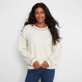 A woman wearing a cream-colored knit sweater with small black speckles and black dotted trim details, paired with blue jeans, against a plain white studio background.