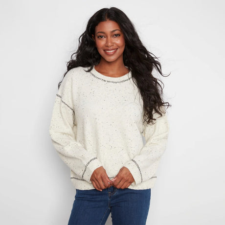 A woman wearing a cream-colored knit sweater with small black speckles and black dotted trim details, paired with blue jeans, against a plain white studio background.
