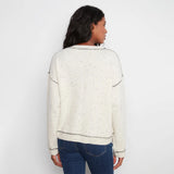 A woman viewed from the back wearing a cream-colored knit sweater with small black speckles and black dotted trim details, paired with blue jeans, against a plain white background.