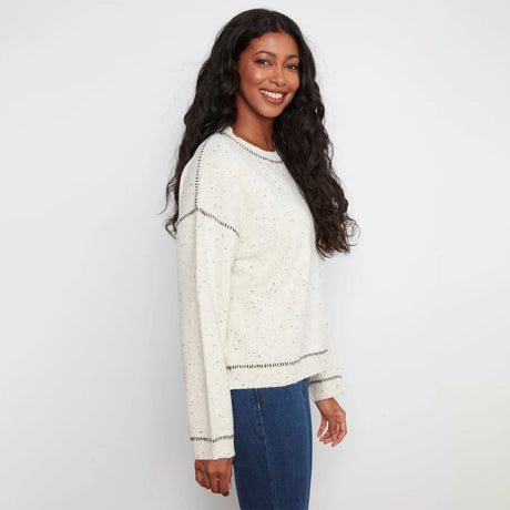 A woman with long, wavy black hair is shown from a side-back angle wearing a cream-colored knit sweater with subtle speckles and dark contrast stitching, paired with blue jeans, against a plain white studio background.