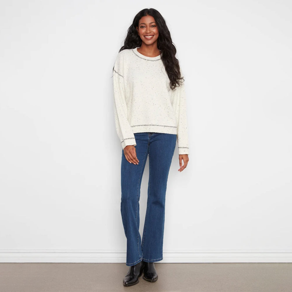A woman stands against a white wall wearing a cream speckled knit sweater, blue flare jeans, and black boots.