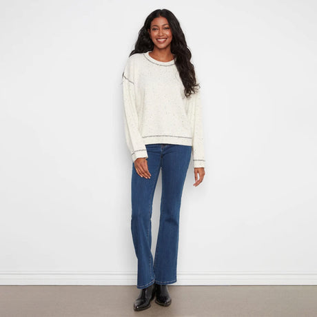 A woman stands against a white wall wearing a cream speckled knit sweater, blue flare jeans, and black boots.