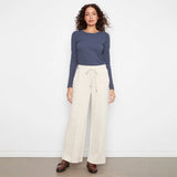 A woman stands against a plain white wall wearing a slate blue long-sleeve top and cream wide-leg drawstring pants, paired with brown loafers.