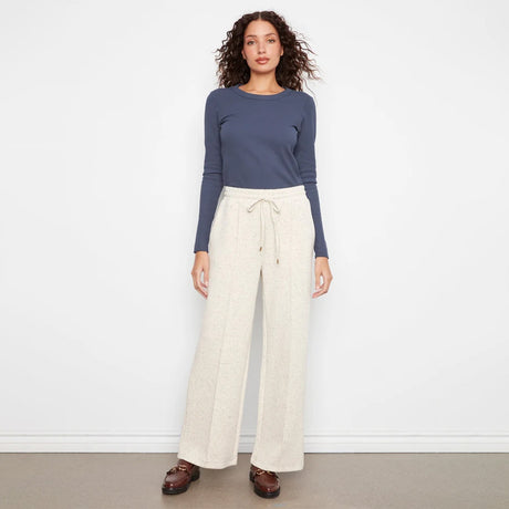 A woman stands against a plain white wall wearing a slate blue long-sleeve top and cream wide-leg drawstring pants, paired with brown loafers.
