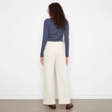 A woman is shown from behind wearing a slate blue long-sleeve top and cream wide-leg trousers, standing against a plain white wall.