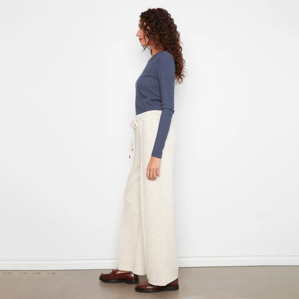 A woman stands in profile wearing a slate blue long-sleeve top, cream wide-leg knit pants, and brown leather shoes against a plain white wall.