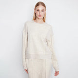 A woman modeling a cream-colored, textured knit crewneck sweater and matching trousers against a plain white studio background.