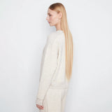 A woman with long blonde hair is shown in profile wearing a textured cream knit sweater and matching knit pants against a plain white studio background.
