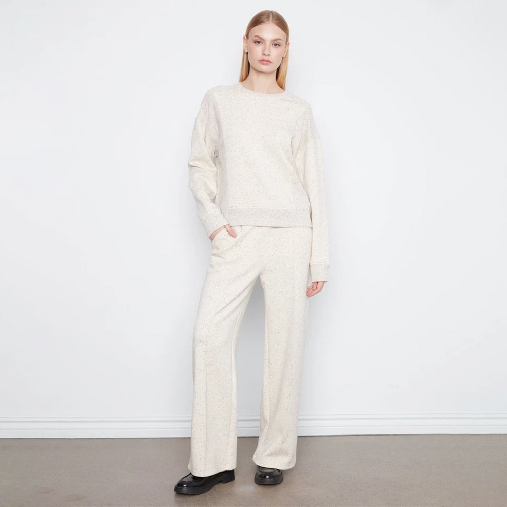 A woman stands against a white wall wearing a matching cream knit sweater and wide-leg trousers set, paired with black shoes.