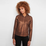 A woman with curly light brown hair wearing a metallic bronze-brown cropped hoodie with black pants, standing against a light gray studio background.