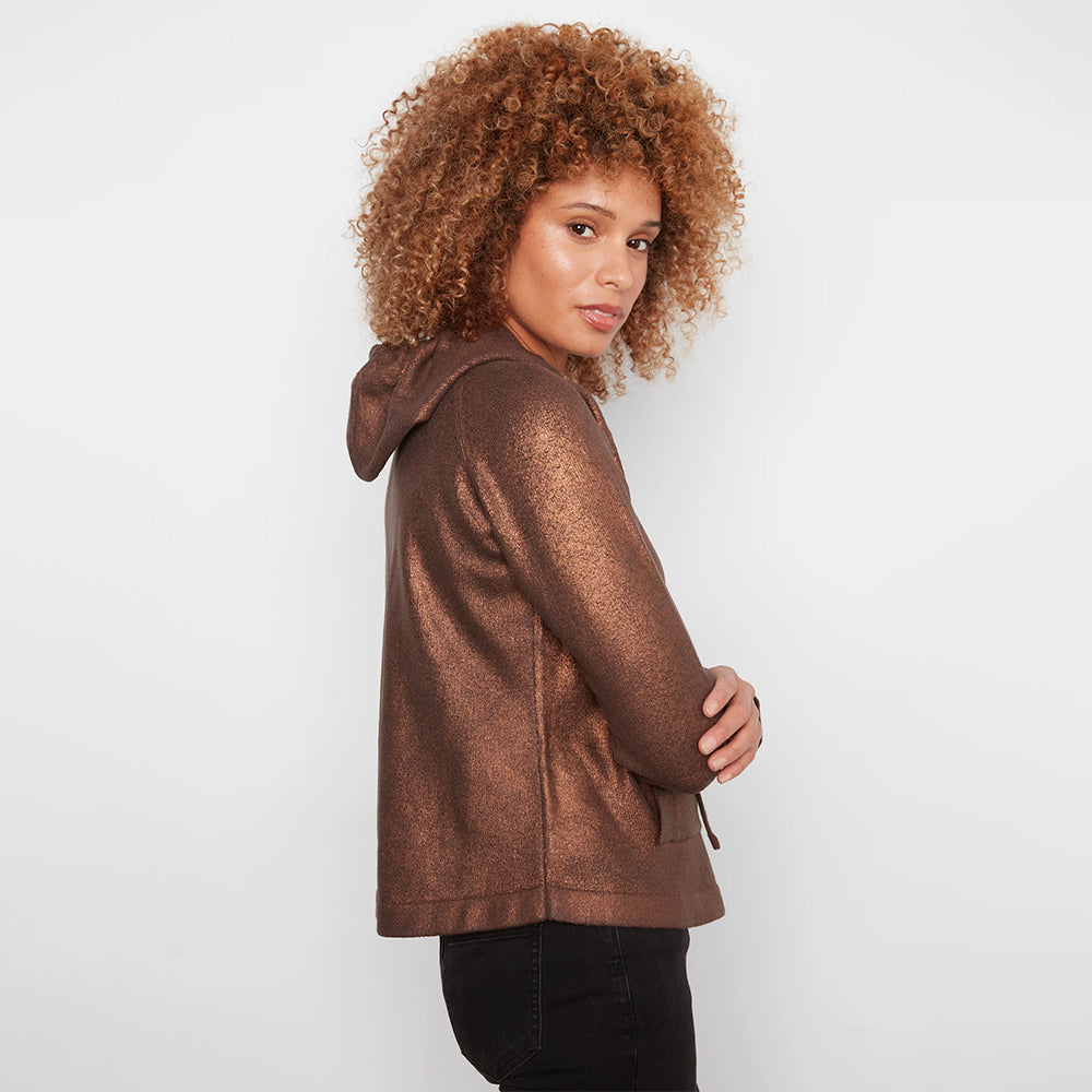 A woman with curly light brown hair is shown from the side/back wearing a metallic bronze-brown hooded sweatshirt and black pants against a plain light gray studio background.